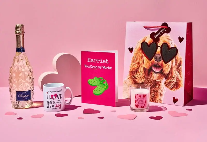 Valentine’s gift display featuring a bottle of sparkling drink, a dog‑themed gift bag, a mug, a bright pink card, and a candle arranged on a pink background with scattered heart decorations.